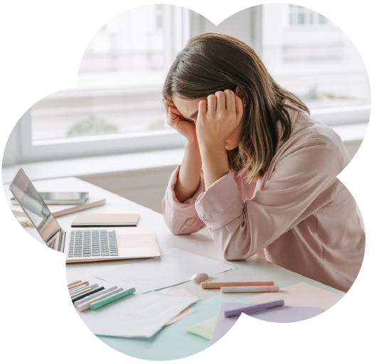 Woman sitting at a desk with her head in her hands, appearing overwhelmed or stressed. The workspace includes a laptop, pastel stationery, markers, and papers scattered across the surface, suggesting creative or work-related frustration.
